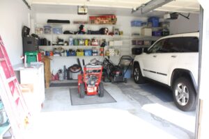 Clean, well-organized residential garage with white walls, storage shelves filled with boxes and supplies, a white SUV parked inside, and a red pressure washer on the concrete floor