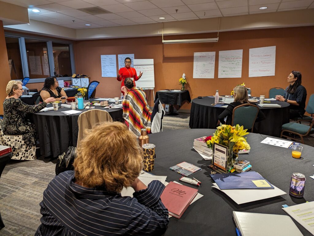 Facilitator leading a workshop session on event marketing for women leaders, with attendees seated at round tables listening and taking notes in a conference room.