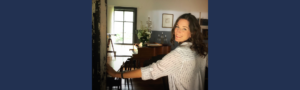 Woman smiling beside a piano in a bright room, representing overcoming obstacles in leadership with calm and resilience